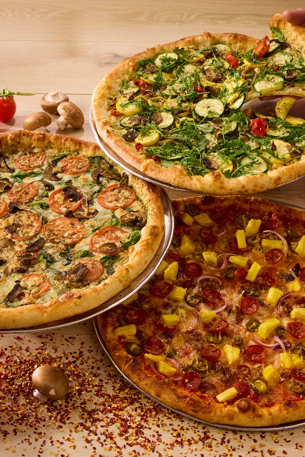 Three colorful pizzas overlap on a wooden table: mushroom and herb, leafy vegetable, and pepper-topped varieties create an appetizing spread.