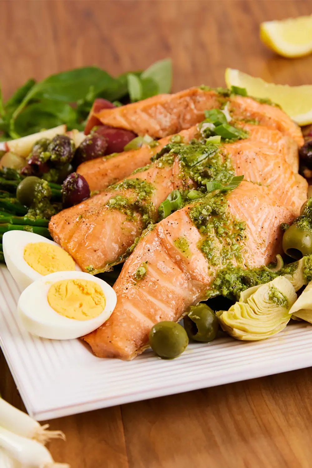 Grilled salmon fillets topped with pesto, asparagus, olives, artichokes, lemon wedges, and sliced hard-boiled eggs on a white platter.