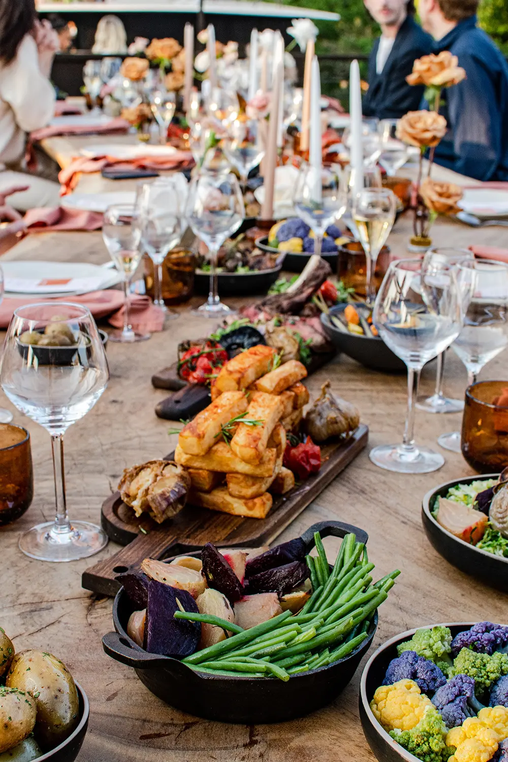 Elegant garden dinner table laden with roasted vegetables, bread, wine glasses, candles, and floral dishes, creating a warm festive mood.