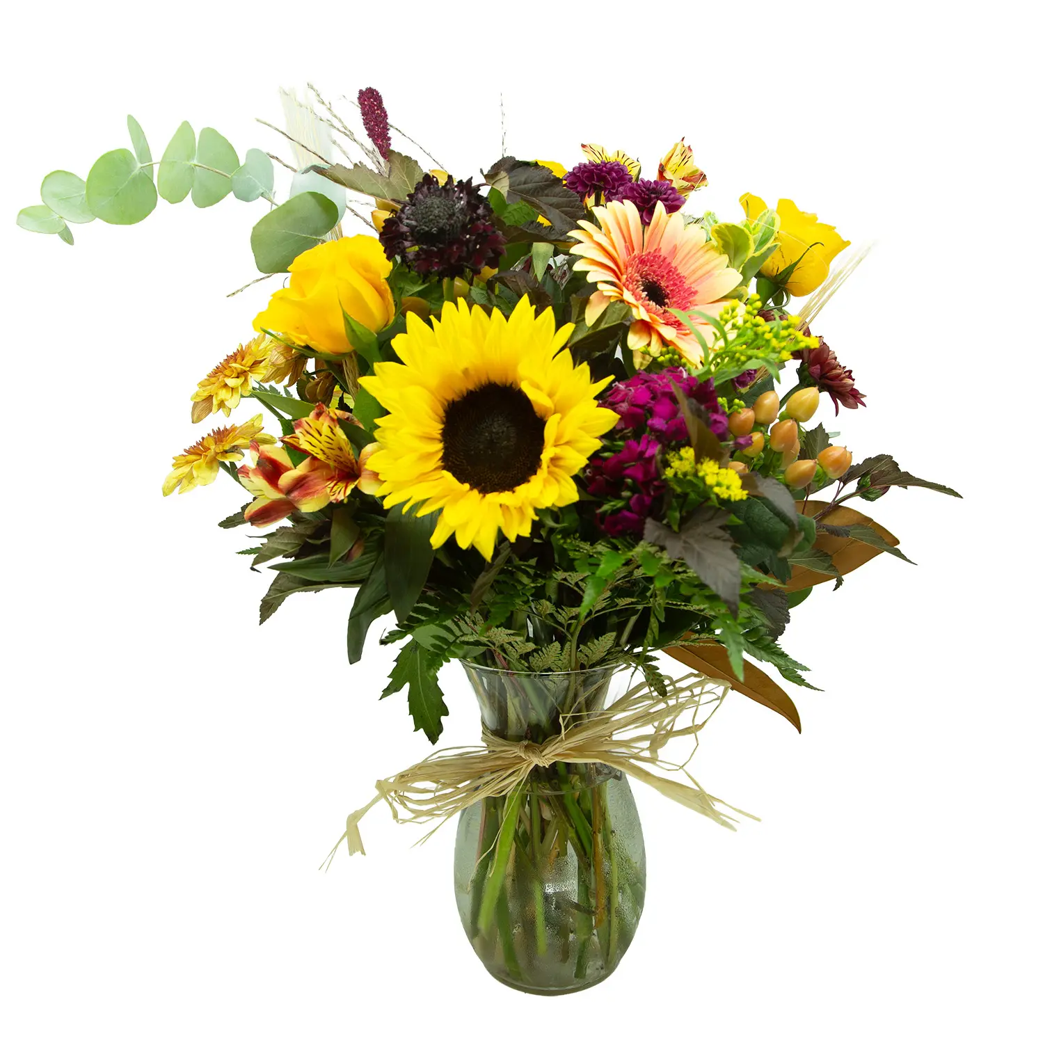 A vibrant mixed bouquet of sunflowers, yellow roses, pink and orange blooms, and greenery sits in a glass vase tied with raffia.