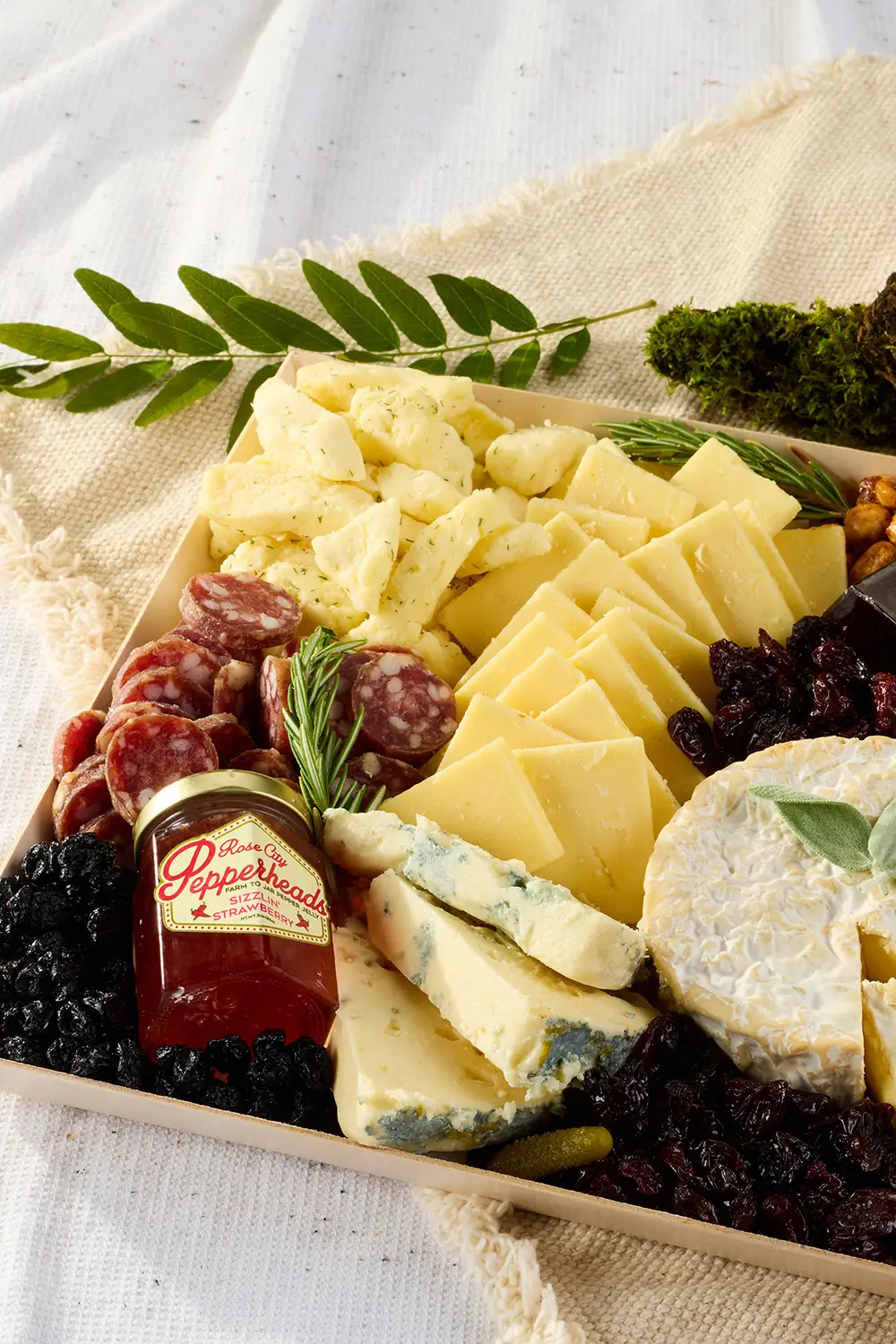 An inviting charcuterie board displays sliced cheeses, salami, pepper jelly, berries, olives, and rosemary on a linen-covered table.