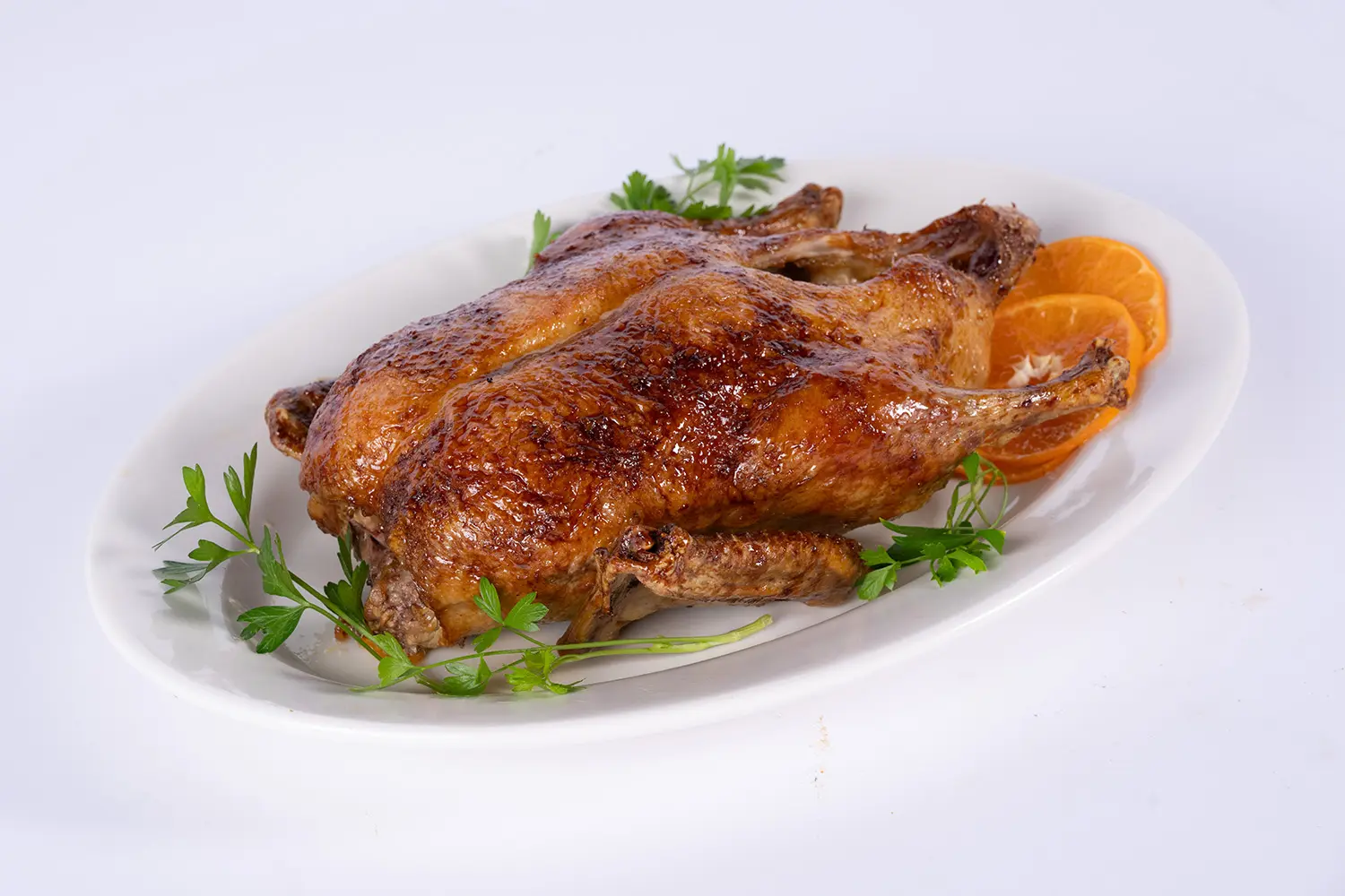 A golden-brown roasted duck rests on a white oval platter, garnished with fresh parsley and orange slices, creating a rich, festive meal presentation.