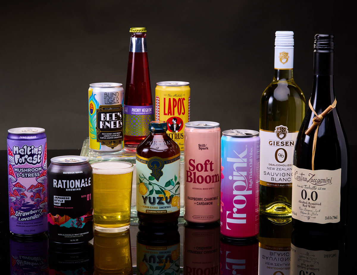 A lineup of colorful zero-proof and alcohol-removed beverages arranged on a glossy black surface, including canned mocktails, botanical sodas, non-alcoholic beers, and alcohol-removed wines. Some of the featured beverages are made in Oregon, and perfect for your Dry January goals.