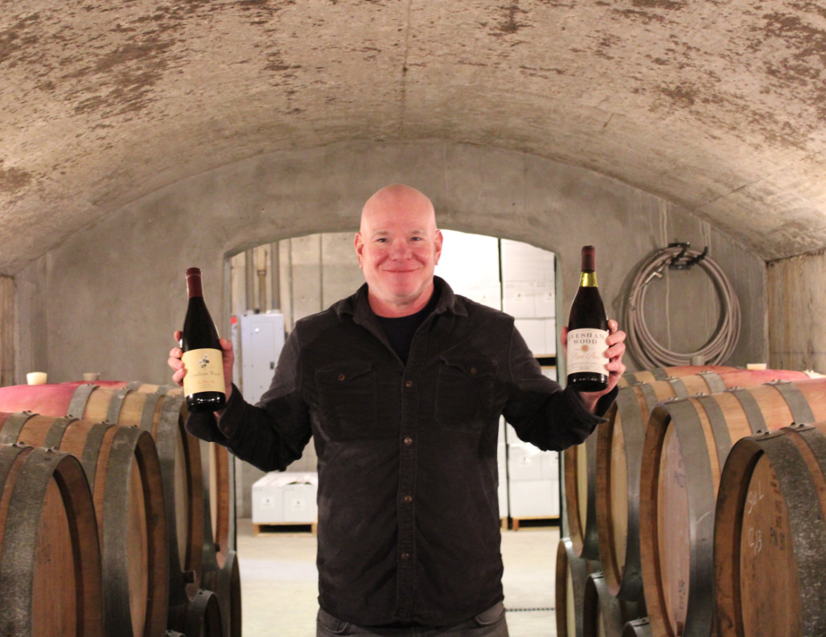 Erin Nuccio stands in a wine cellar at Evesham Wood, smiling and holding two bottles of wine, with rows of oak barrels lining both sides behind him.