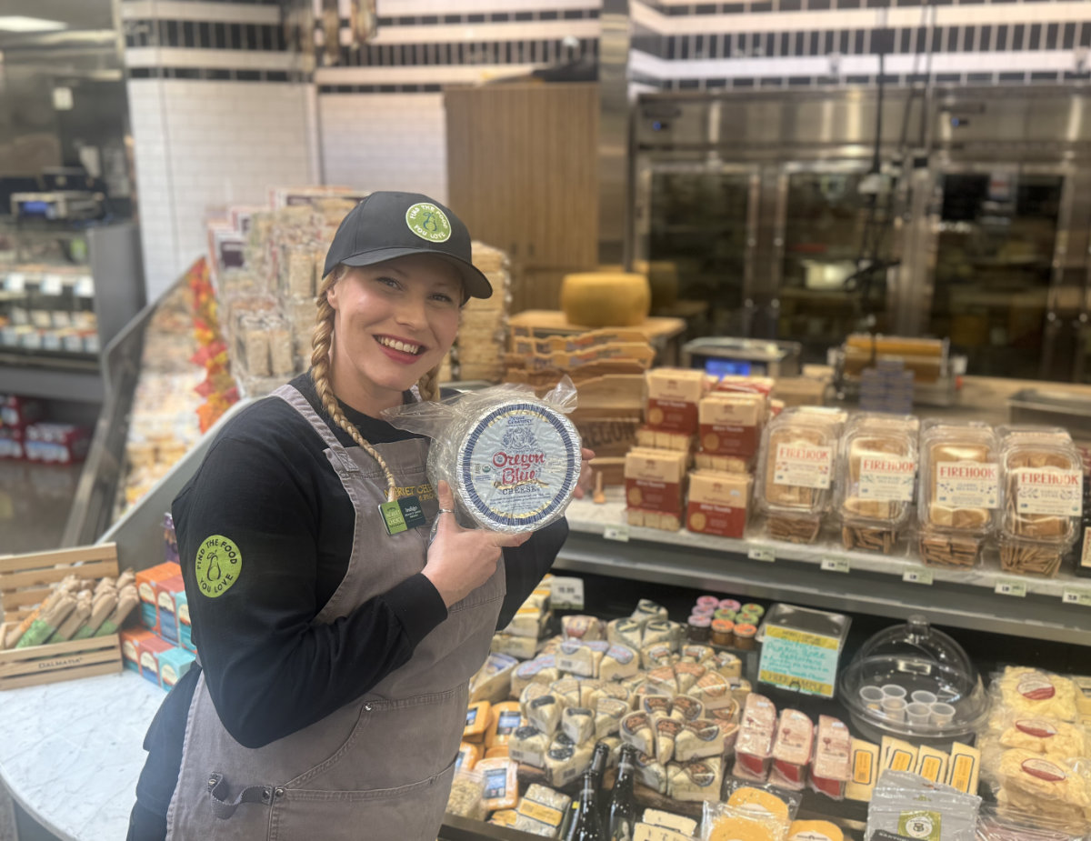 Smiling Market of Choice Cheesemonger Indigo stands behind the cheese counter, holding a wheel of Oregon Blue cheese, with a full display of specialty cheeses and accompaniments in the background.