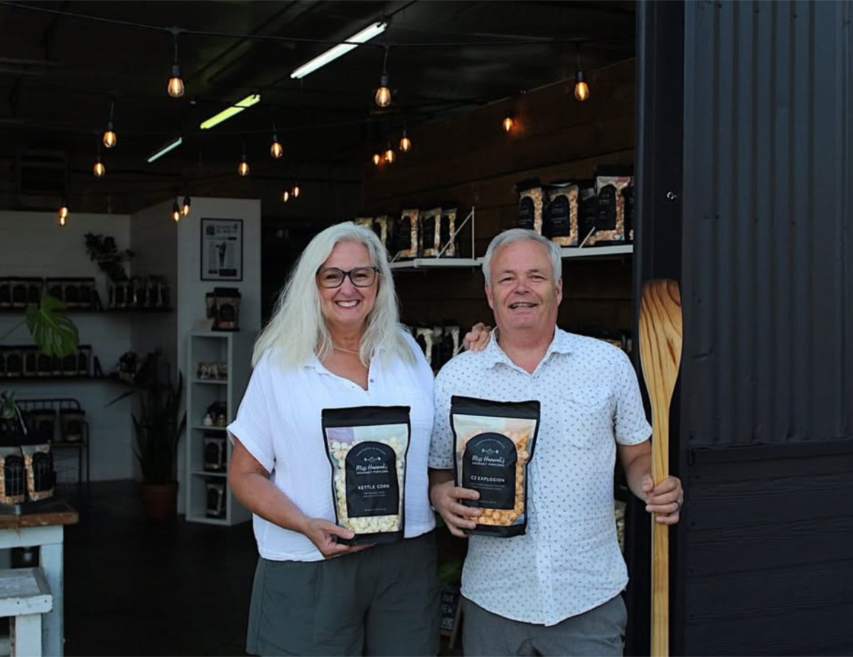 Two owners of Hannah’s Gourmet Popcorn stand smiling in the doorway of a small shop, holding bags of Gourmet Popcorn, with shelves of packaged popcorn visible behind them.