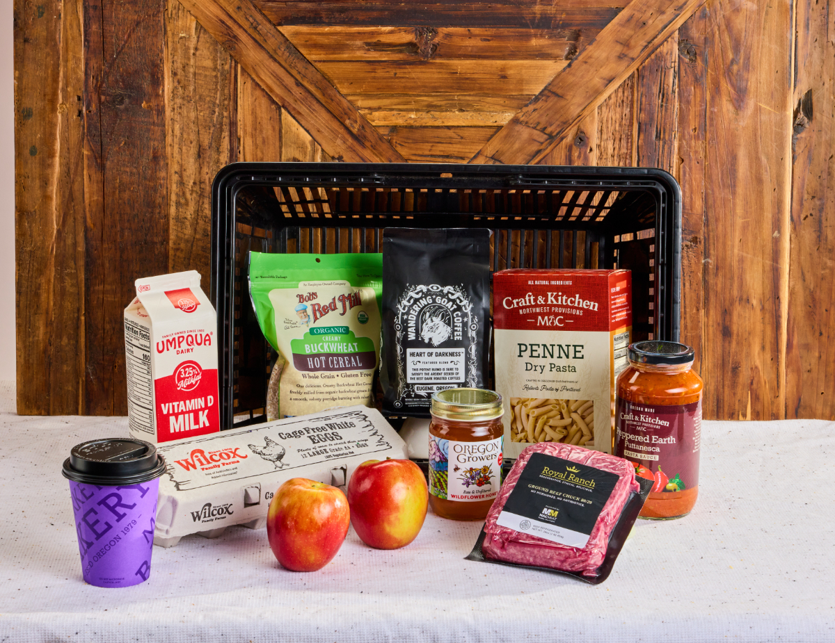 Shopping basket filled with grocery staples — Umpqua milk, Wilcox eggs, Bob's Red Mill buckwheat cereal, Wandering Goat coffee, Craft & Kitchen penne and puttanesca sauce, Oregon Growers honey, Royal Ranch ground beef, apples, and a bakery coffee cup.