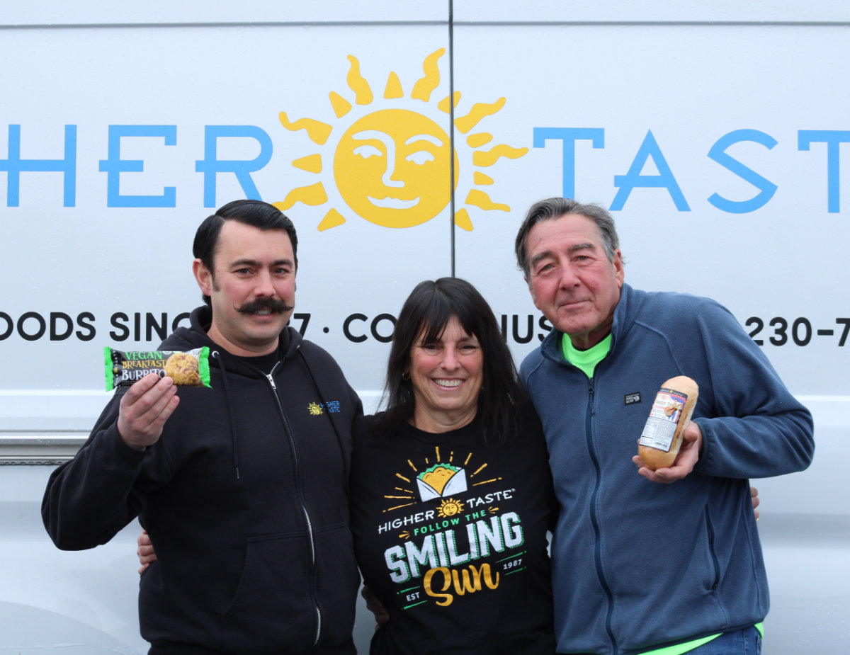 Three members of the Higher Taste team stand smiling in front of a branded delivery truck, holding plant-based products including a vegan breakfast burrito and a packaged entrée, showcasing their Oregon-made foods.