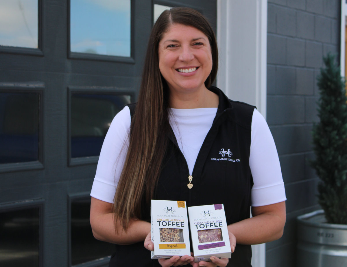 Randy Holm standing outside a production space holding two boxes of Oregon hazelnut toffee, smiling and wearing a black vest with the Holm Made Toffee Co. logo