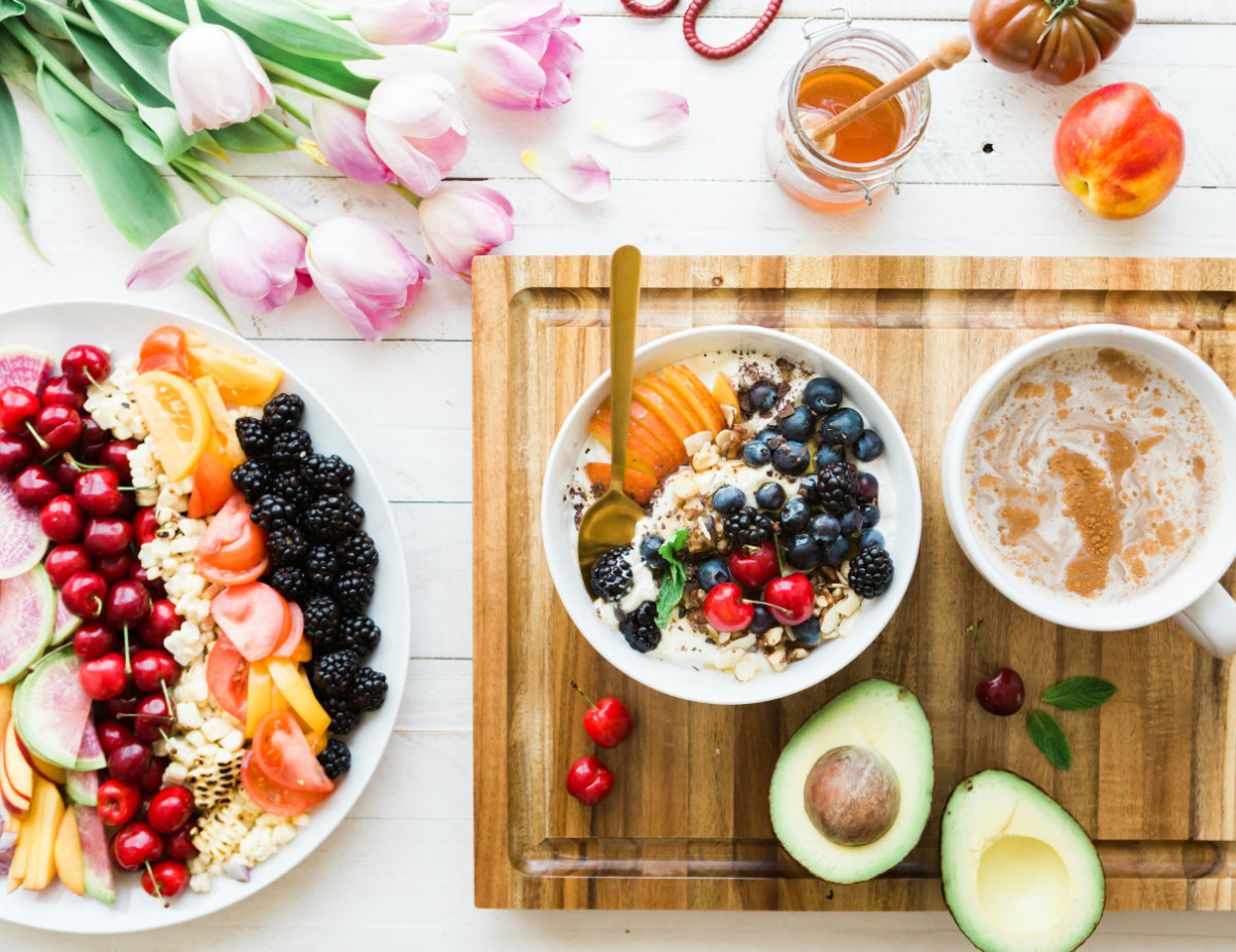 Overhead view of a colorful healthy breakfast spread featuring yogurt topped with berries and nuts, fresh fruit salad, avocado halves, honey, coffee and pink tulips on a white table.