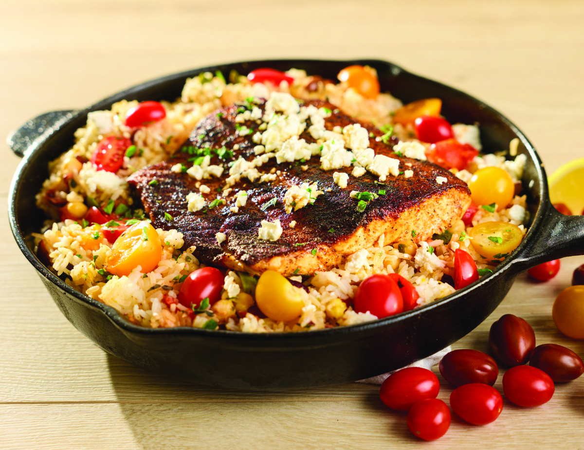 Mediterranean Salmon Skillet — seared salmon fillet seasoned with smoked paprika over Greek-style fried rice with chickpeas, Kalamata and green olives, multicolored cherry tomatoes, feta crumbles, and fresh herbs in a cast iron pan, served with a lemon wedge.