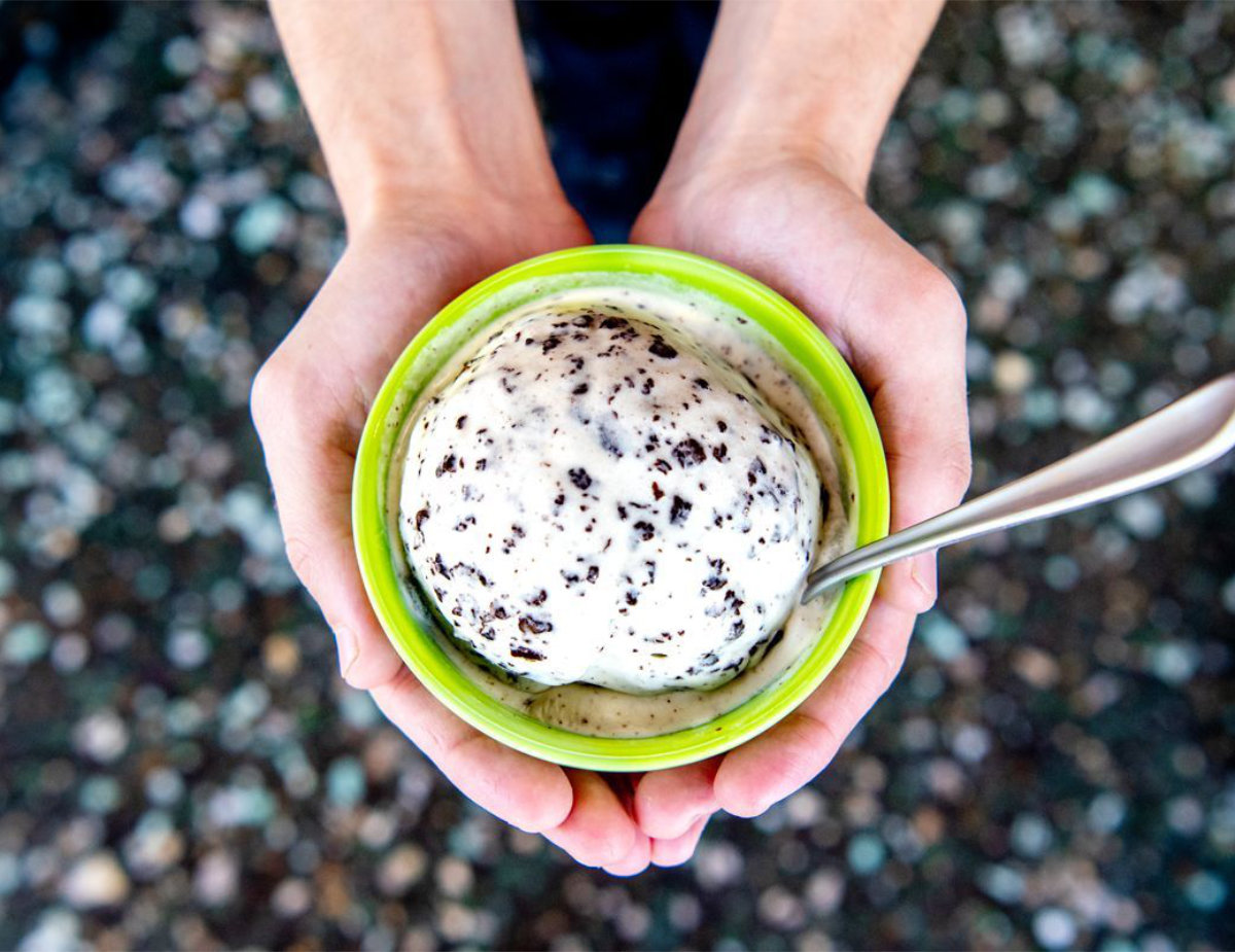 Overhead view of two hands holding a small green bowl of cookies-and-cream ice cream with a spoon, set against a blurred pebbled background.