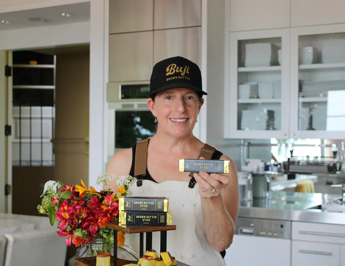 Smiling woman in a kitchen wears a black cap and apron, holding a product box beside colorful flowers in a warm promotional scene.