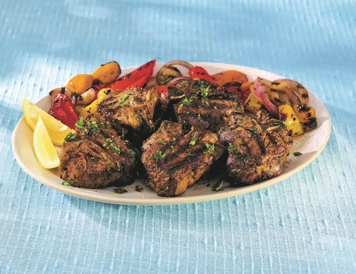 Grilled lamb chops with charred vegetables, herbs, and lemon wedges arranged on a white platter, evoking a hearty Mediterranean meal.
