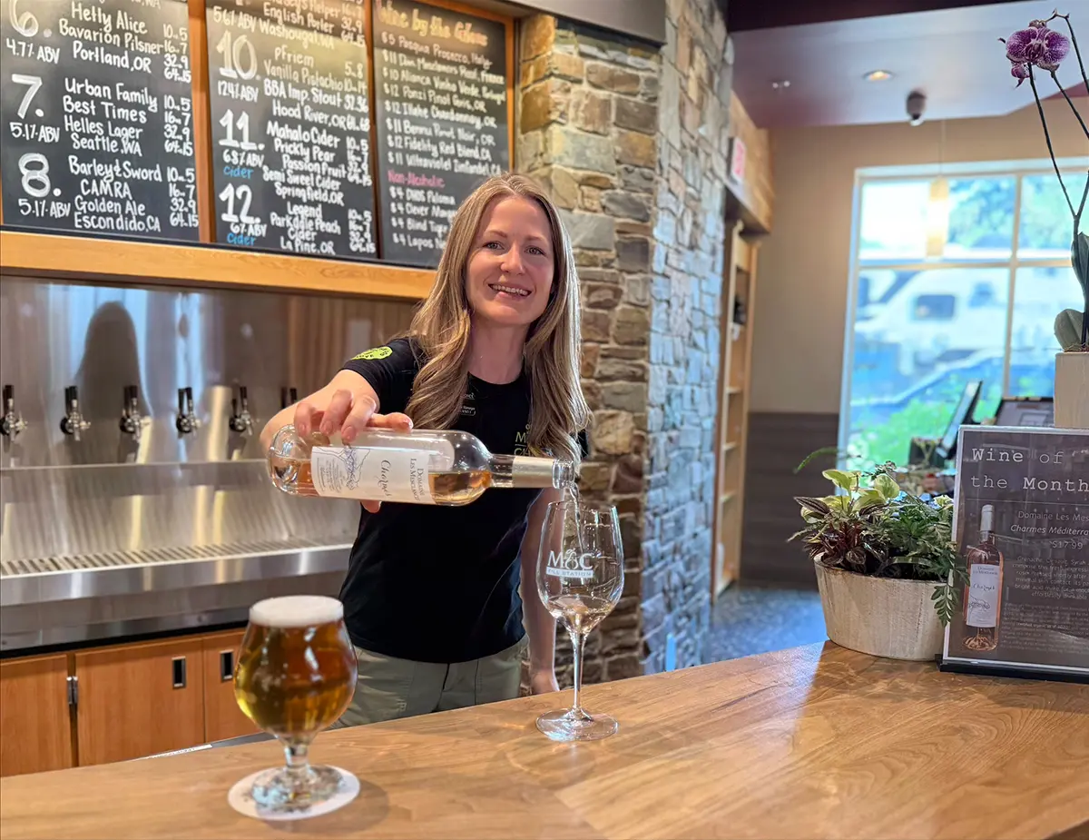 Smiling brewery server pours beer into a stemmed glass at a wooden bar, with taps, chalkboard menu, stone wall, and warm taproom décor.