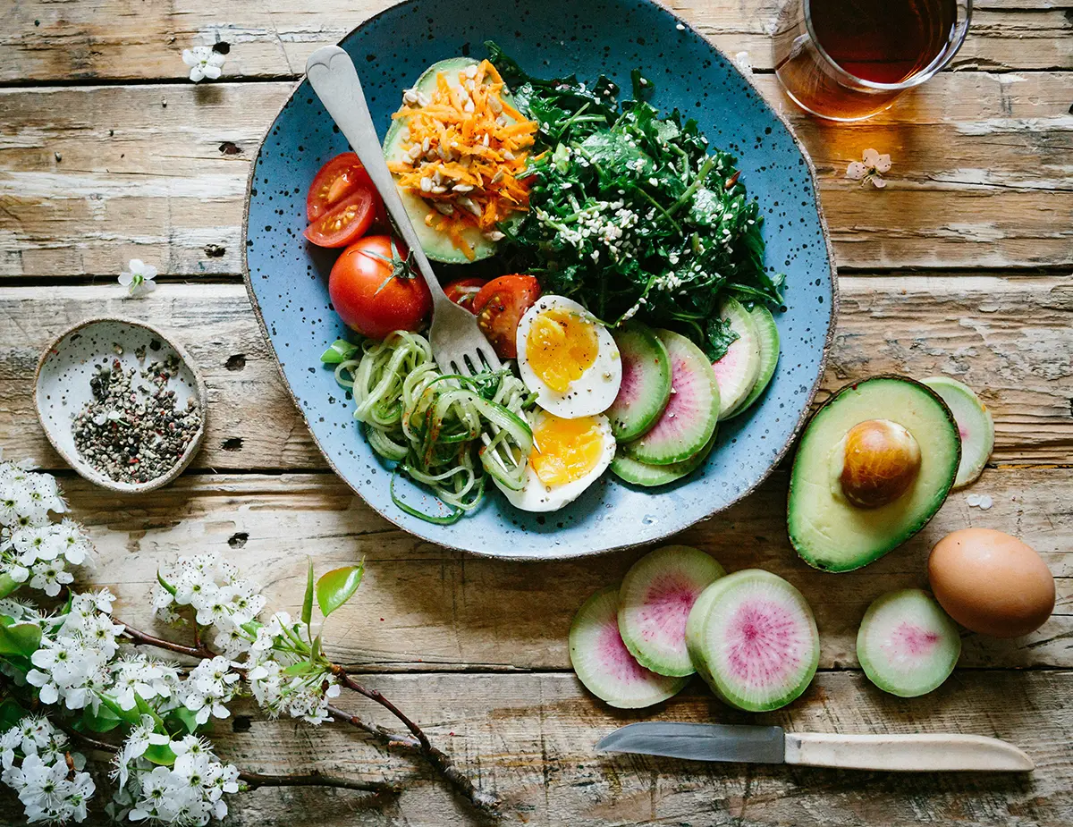 Colorful healthy salad bowl with greens, carrots, tomatoes, zucchini noodles, radishes, avocado, and boiled eggs on a rustic wooden table.